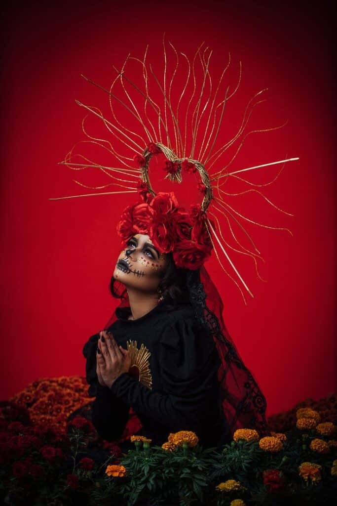 Person in traditional Day of the Dead makeup and ornate headpiece surrounded by vibrant flowers against a red background.