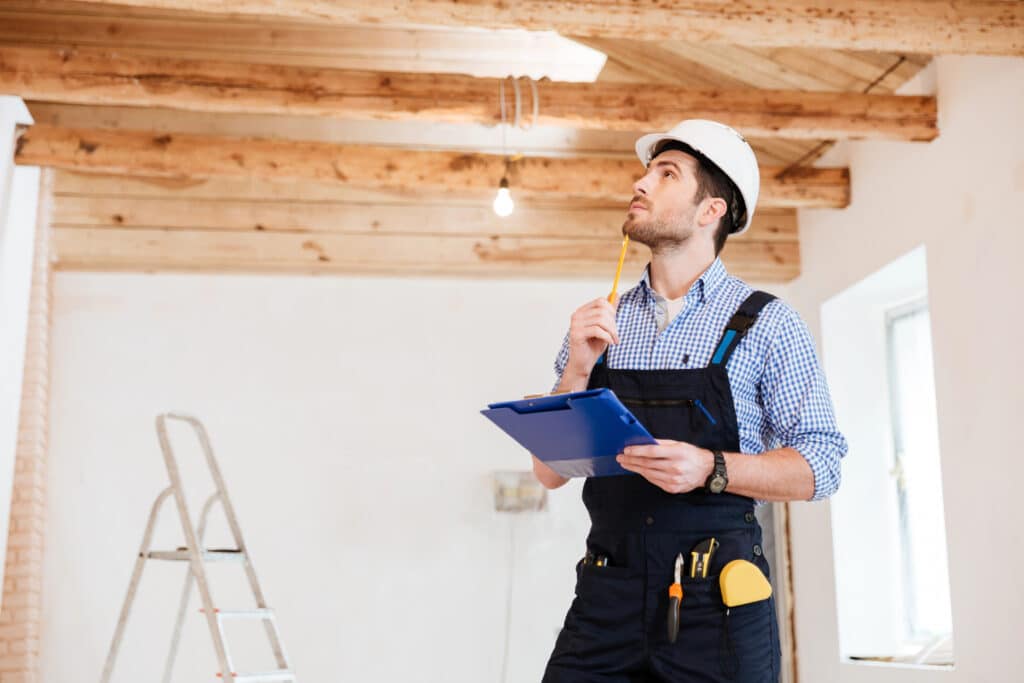 Construction worker in hard hat inspecting ceiling, holding clipboard and pencil, ladder in background.