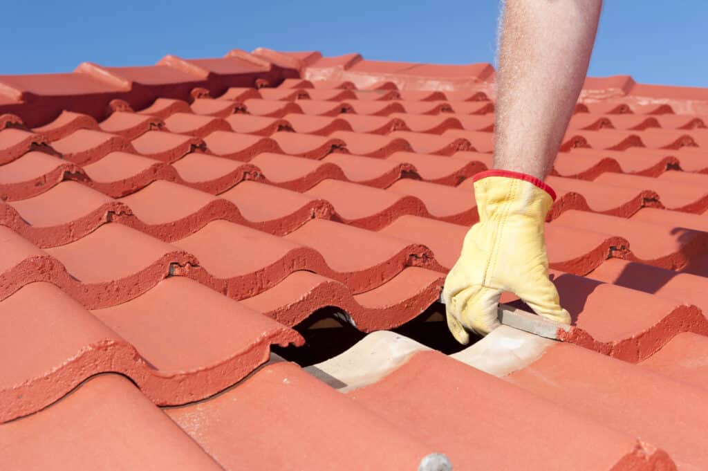 Red roof tile repair in progress with gloved hand adjusting tiles under clear blue sky.