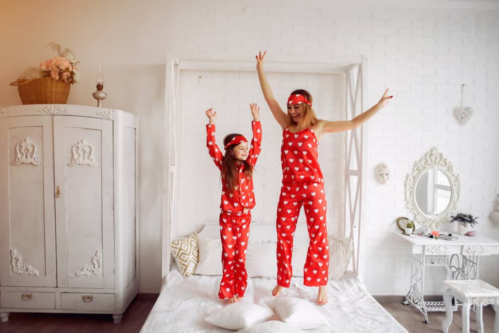 Mother and daughter in red heart pajamas joyfully jumping on bed in bright bedroom.