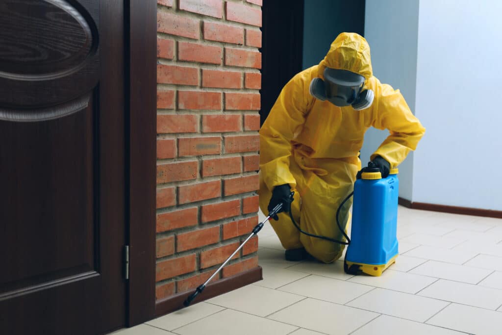 Worker in yellow hazmat suit spraying pesticides indoors for pest control, near brick wall and wooden door.