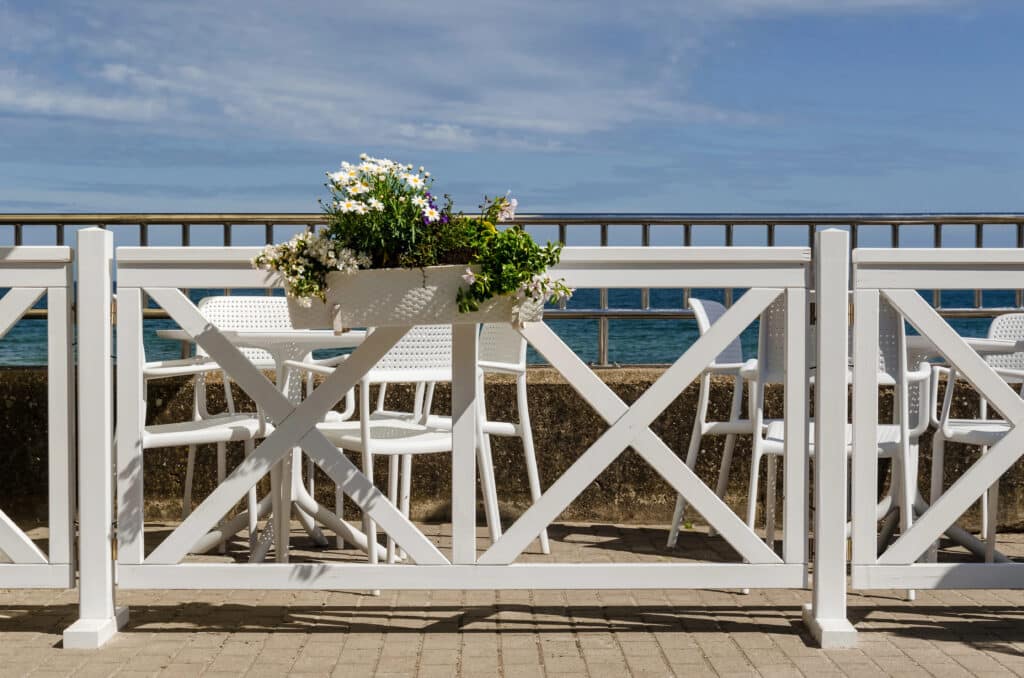 Depositphotos_387122148_XL White patio chairs and flowers on a seaside deck, clear blue sky backdrop. | Sky Rye Design White patio chairs and flowers on a seaside deck, clear blue sky backdrop.
