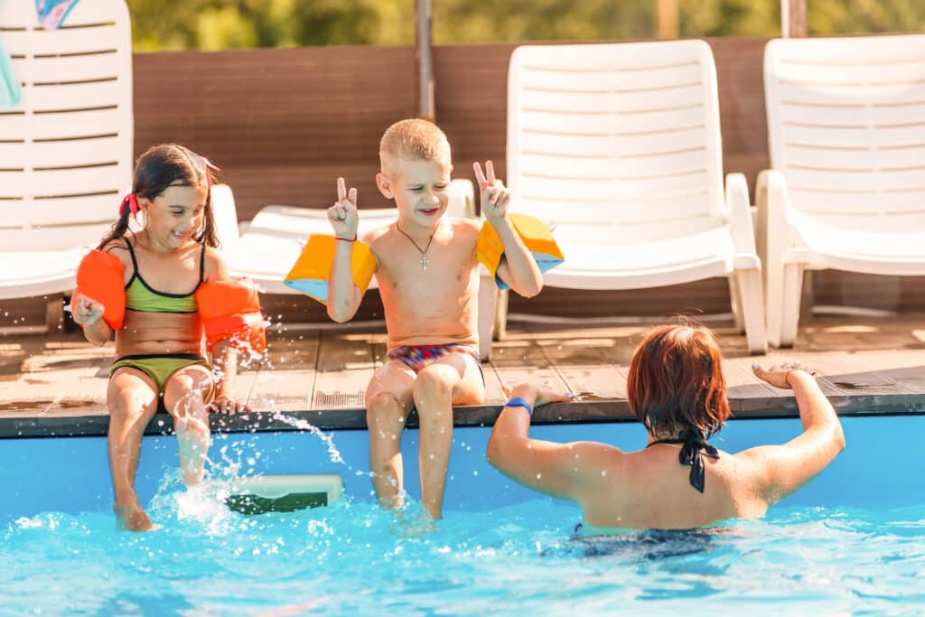 Kids in swim gear play by the poolside with an adult in the water. Bright sunny day at a poolside deck.