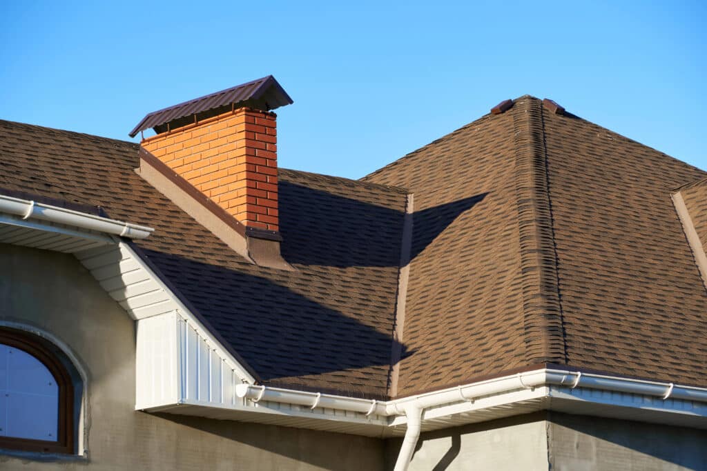 Brown shingle roof with chimney and gutters against blue sky. Ideal for roofing and construction themes.