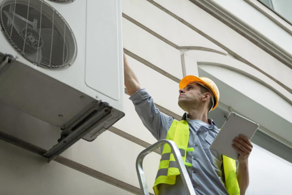 Depositphotos_483063204_XL Technician on ladder inspecting air conditioning unit on building exterior with tablet and safety gear. | Sky Rye Design Technician on ladder inspecting air conditioning unit on building exterior with tablet and safety gear.