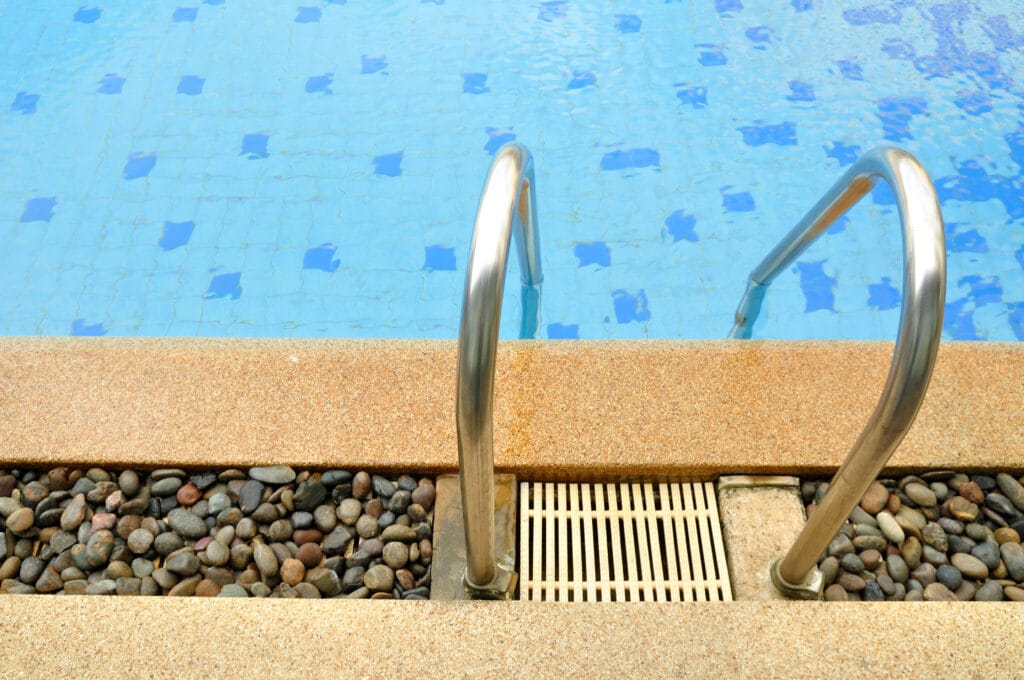 Poolside ladder with stones and tiles, leading into clear blue water. Ideal for summer relaxation.