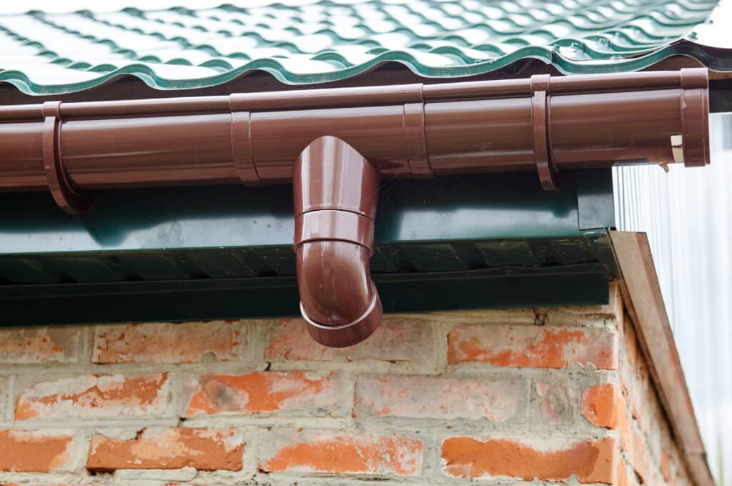 Close-up of a brown gutter and downspout on a building with a green metal roof and brick wall background.