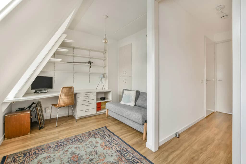 Minimalist home office with desk, chair, shelves, and sofa in an attic room, featuring light wood flooring.