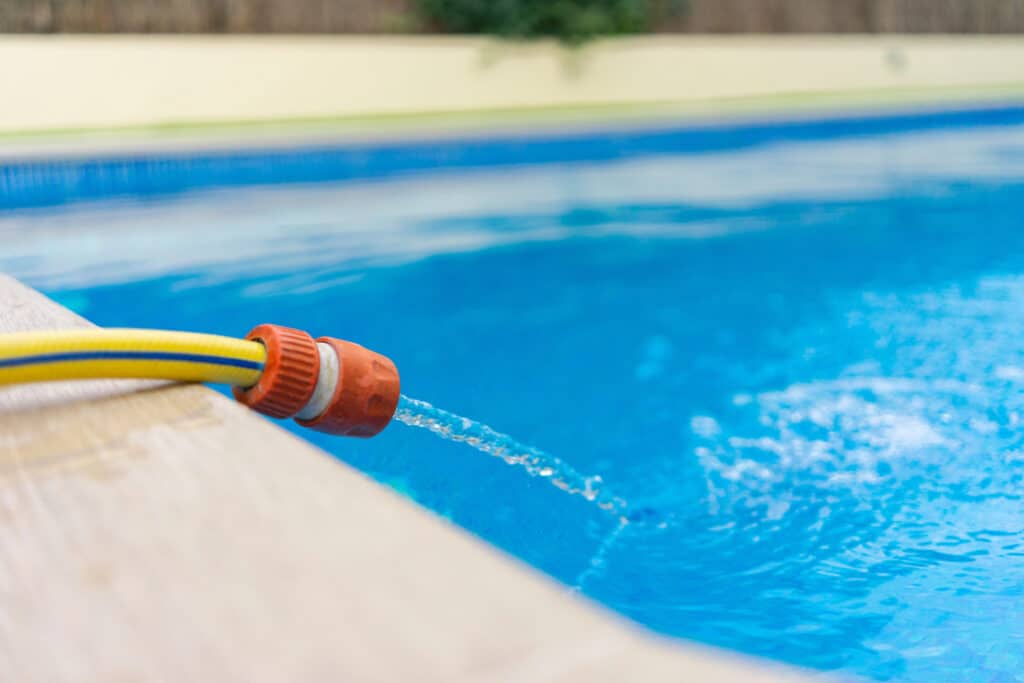 Garden hose filling a swimming pool with clear blue water on a sunny day.