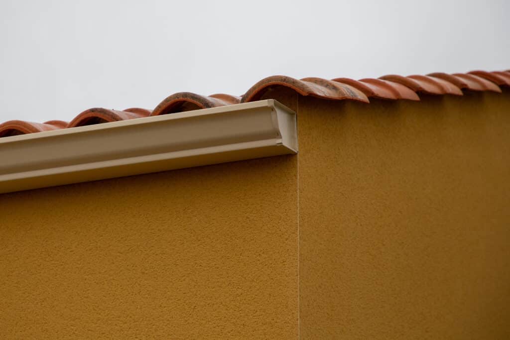 Terracotta tiles and stucco wall with beige metal gutter under a cloudy sky.