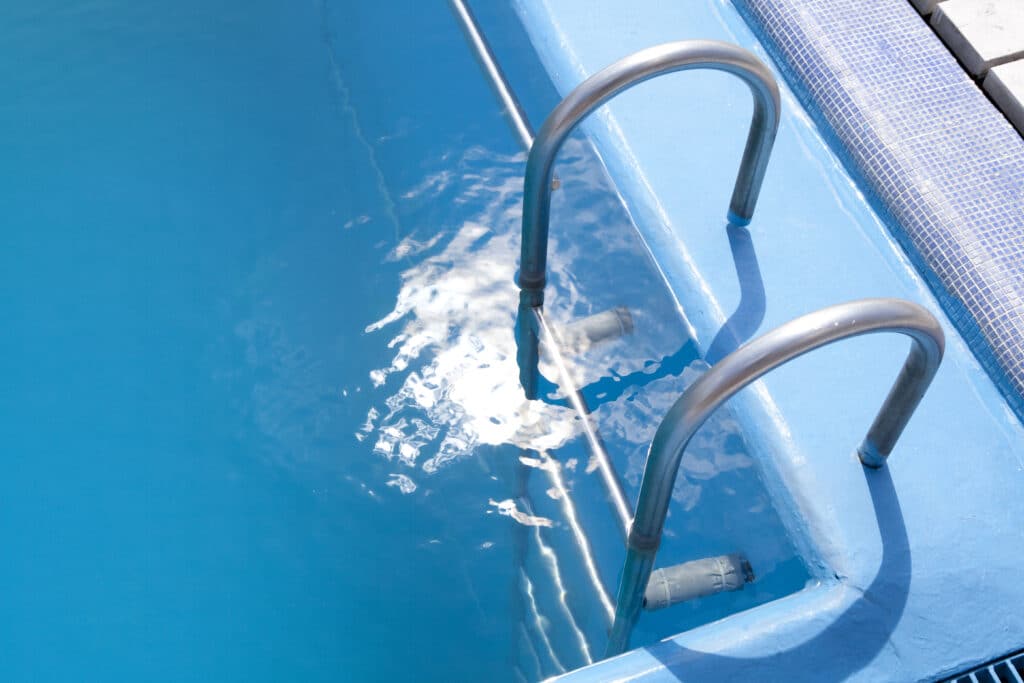Shiny metal pool ladder leading into clear blue water of a swimming pool with sunny reflections.