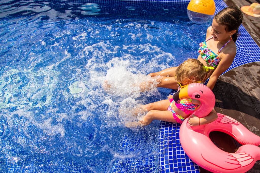 Children playing in a pool with a pink flamingo float, enjoying a sunny day, while splashing water around.