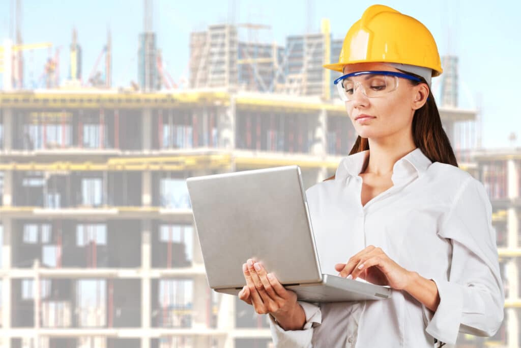 Female engineer in hard hat using laptop at construction site, building in background.