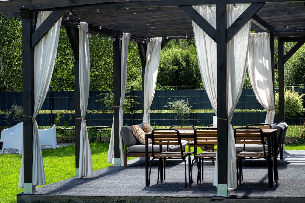 The terrace behind the house is made of beams the roof is covered with polycarbonate Outdoor pergola with dining set, cozy seating, and draped curtains in a lush garden setting. | Sky Rye Design Outdoor pergola with dining set, cozy seating, and draped curtains in a lush garden setting.