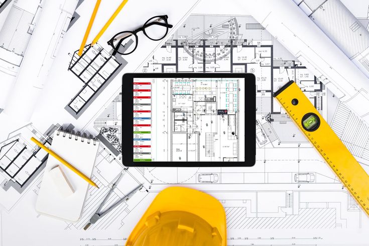 Tablet displaying architectural plans on desk with blueprints, notepad, glasses, and yellow construction tools.