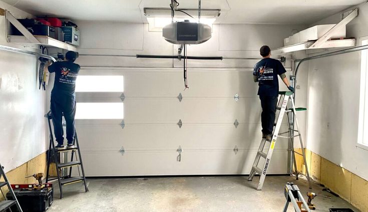 Technicians installing storage shelves in a garage with a white door, using ladders for access and tools on the floor.