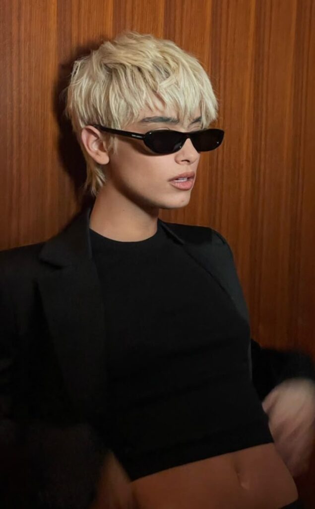 Person with short blonde hair and sunglasses wearing a black outfit against a wooden background.