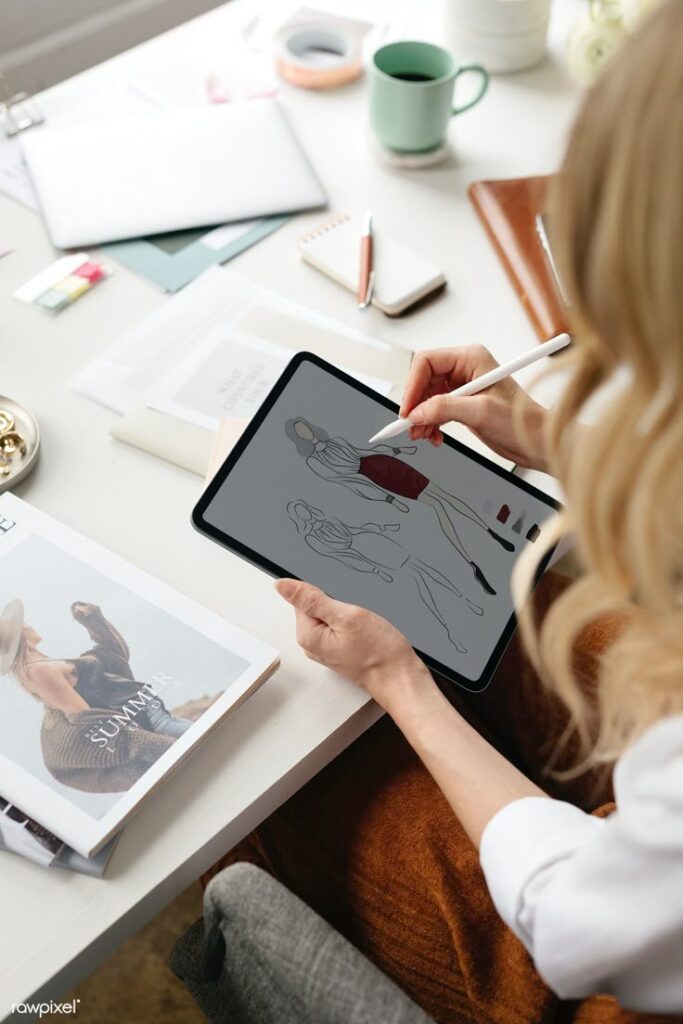 Fashion design illustration on tablet, designer sketching outfit at a creative workspace with books and coffee.