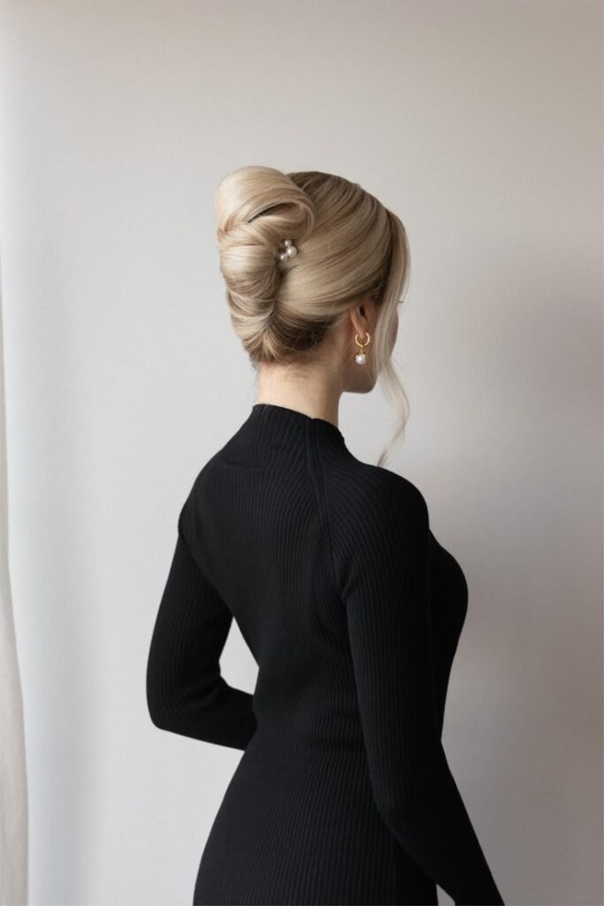 Elegant blonde updo hairstyle with pearl accessories, woman in black dress from behind.