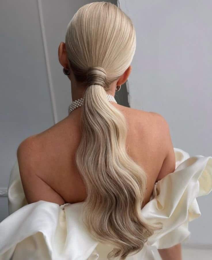 Elegant blonde ponytail with soft waves, styled for a formal event. Woman wearing white off-shoulder gown and pearl choker.