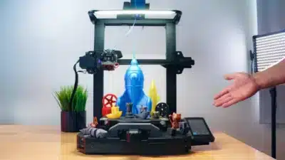 3D printer showcasing various colorful models, including a blue rocket and yellow bust, on a wooden table.