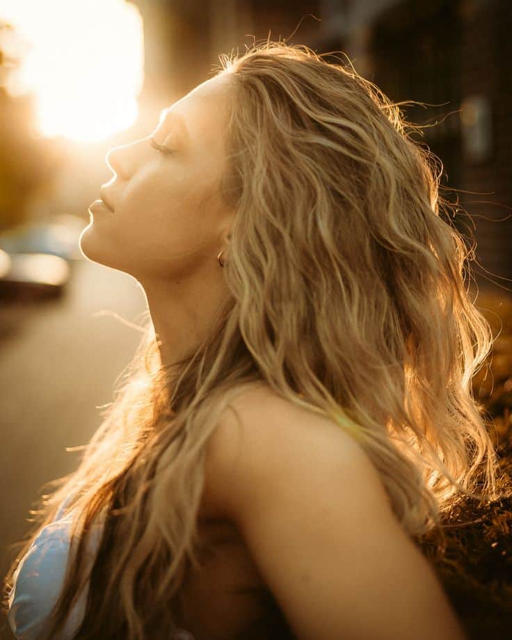 Woman basks in warm sunset glow, eyes closed in peaceful enjoyment. Sunlight highlights her flowing blonde hair.
