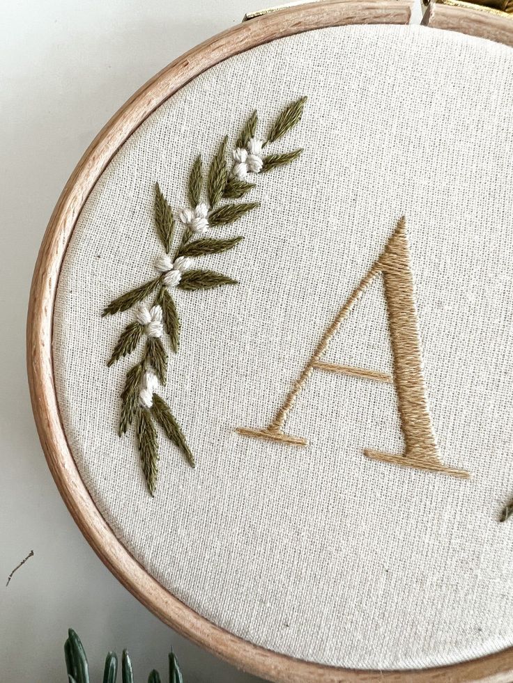 Embroidered letter A with floral design on fabric hoop, showcasing elegant needlework and natural elements.