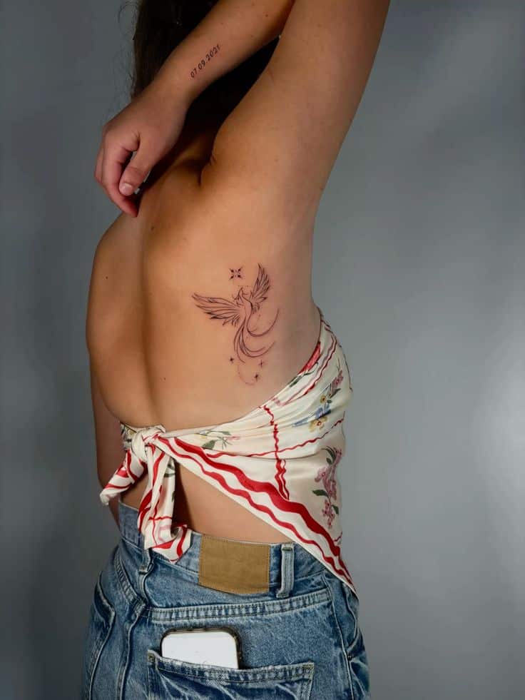 Person with a phoenix tattoo on their side, wearing a floral top and jeans, with a phone in the back pocket.
