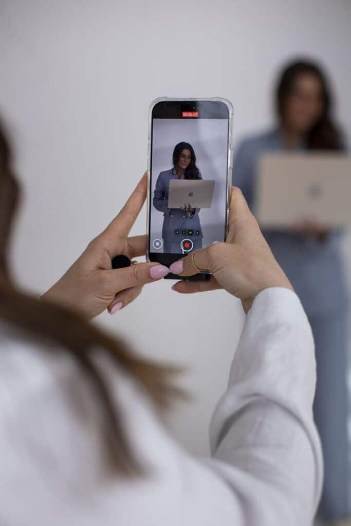 Person recording a woman with a laptop using a smartphone in a bright, minimalist setting.