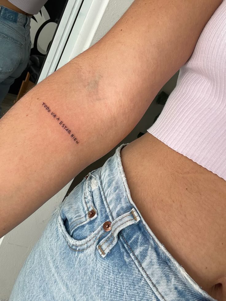 Side view of a person's arm with the tattoo TODO VA A ESTAR BIEN above the elbow, wearing a pink top.