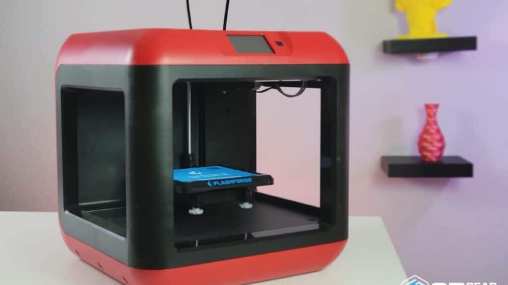 Red 3D printer on a table with shelves in the background displaying a yellow bust and a red vase.