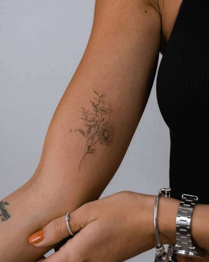Delicate floral tattoo featuring daisies on a woman's forearm in a black sleeveless top and silver bracelet.
