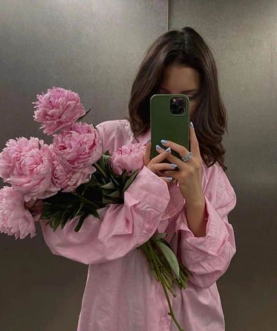 Fotos que debes sacarte con las flores que te regalaste a ti misma Woman in pink shirt takes mirror selfie holding pink peonies, using a smartphone to capture the flowers' beauty. | Sky Rye Design Woman in pink shirt takes mirror selfie holding pink peonies, using a smartphone to capture the flowers' beauty.