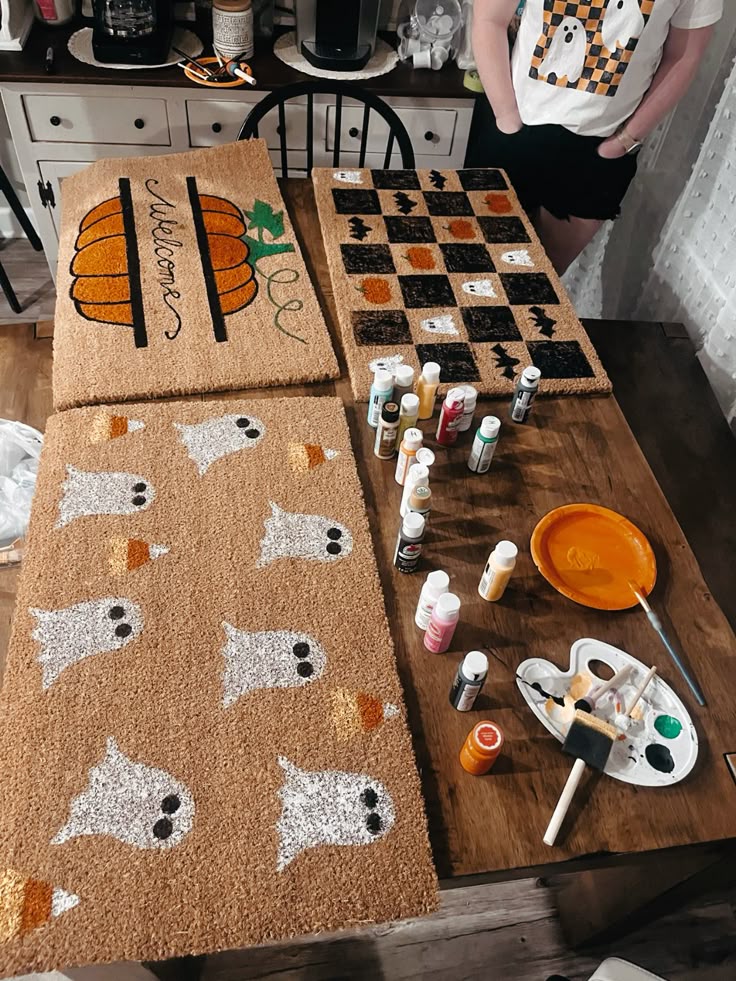 Cozy Halloween-themed doormats with ghosts, bats, and pumpkins on a table with paint supplies.