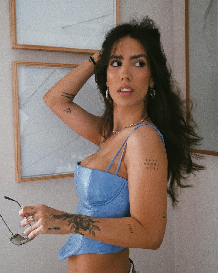 Woman with tattoos in a blue top poses with sunglasses in hand, framed art in the background.