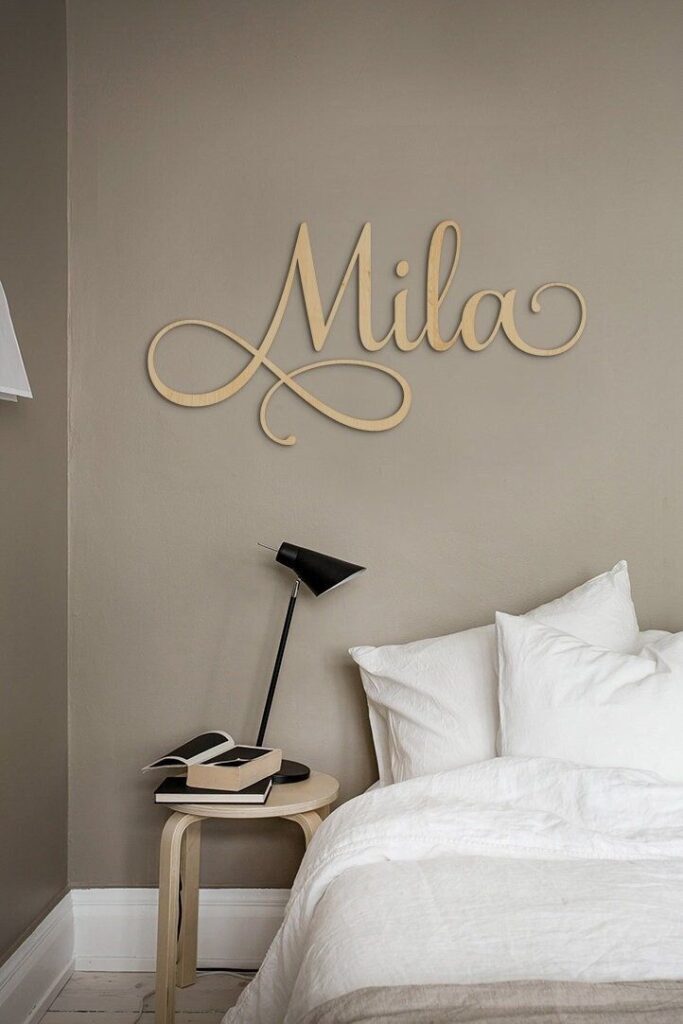 Cozy beige bedroom with white bedding, wall art of the name Mila, a wooden side table, and a black lamp.