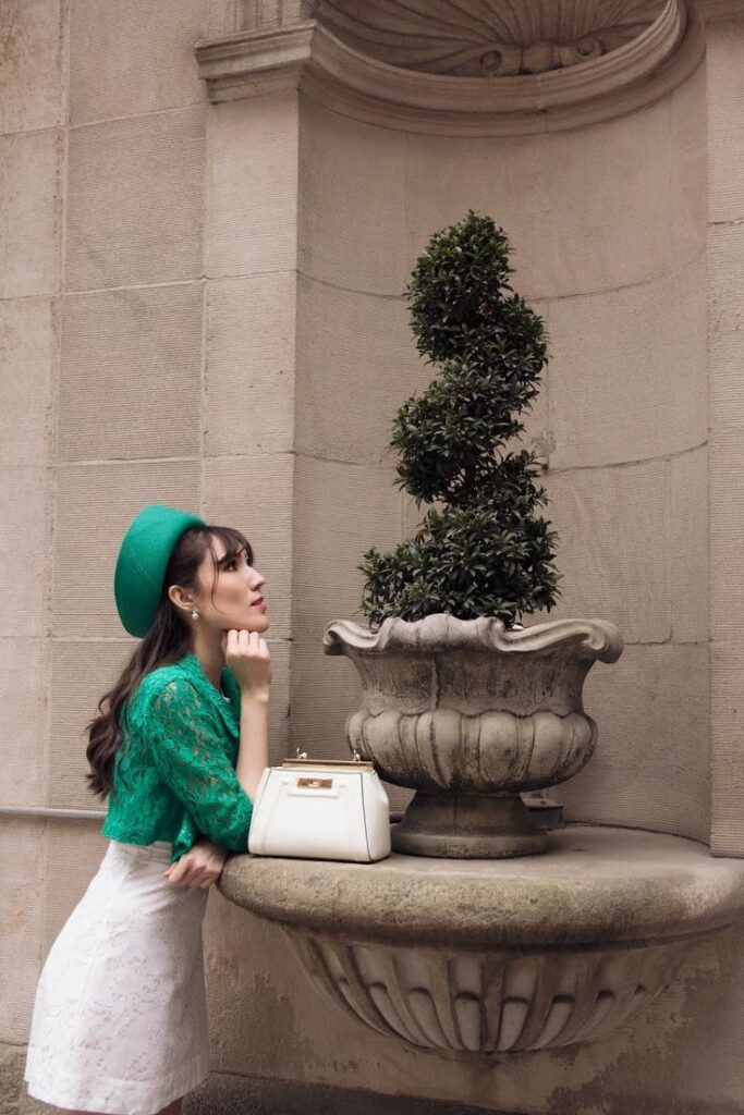Woman in green outfit and hat admires topiary in ornate stone planter, white handbag placed on ledge.