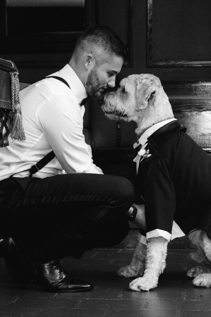 Man in formal attire kneels to lovingly touch noses with a dog dressed in a tuxedo, showcasing a touching bond.