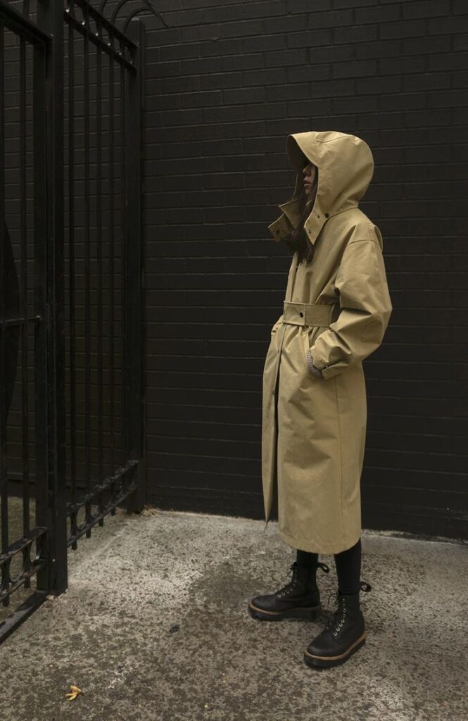 Person in a hooded tan trench coat and boots stands by a black brick wall, exuding urban style and mystery.