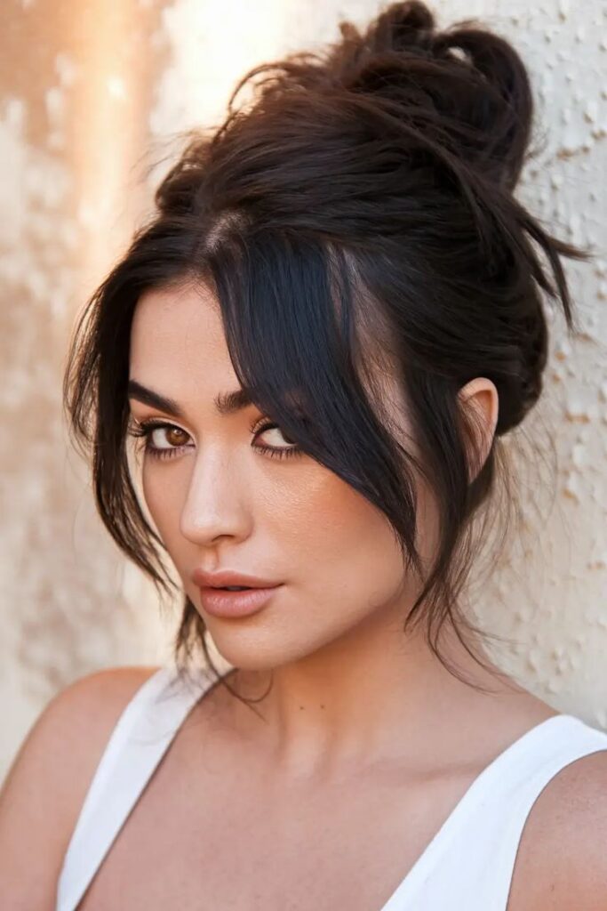 Portrait of a woman with dark hair in a bun, wearing a white top against a textured wall, looking into the distance.