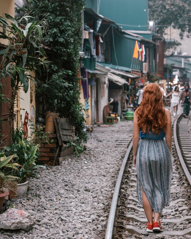 Woman walking on train tracks in a vibrant, colorful street with flags and plants, creating a rustic urban scene.