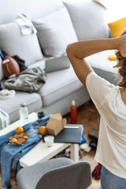 Person overwhelmed by cluttered living room with scattered clothes, food containers, and a laptop on the sofa.