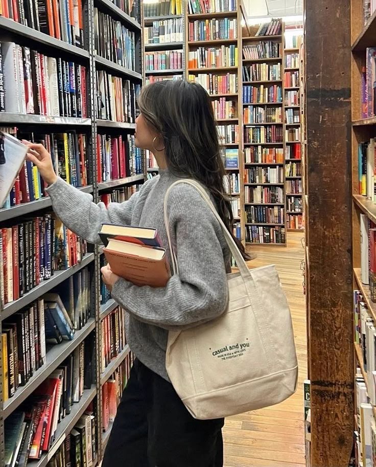 Person browsing books in a library, holding a tote bag and books, exploring shelves filled with colorful spines.