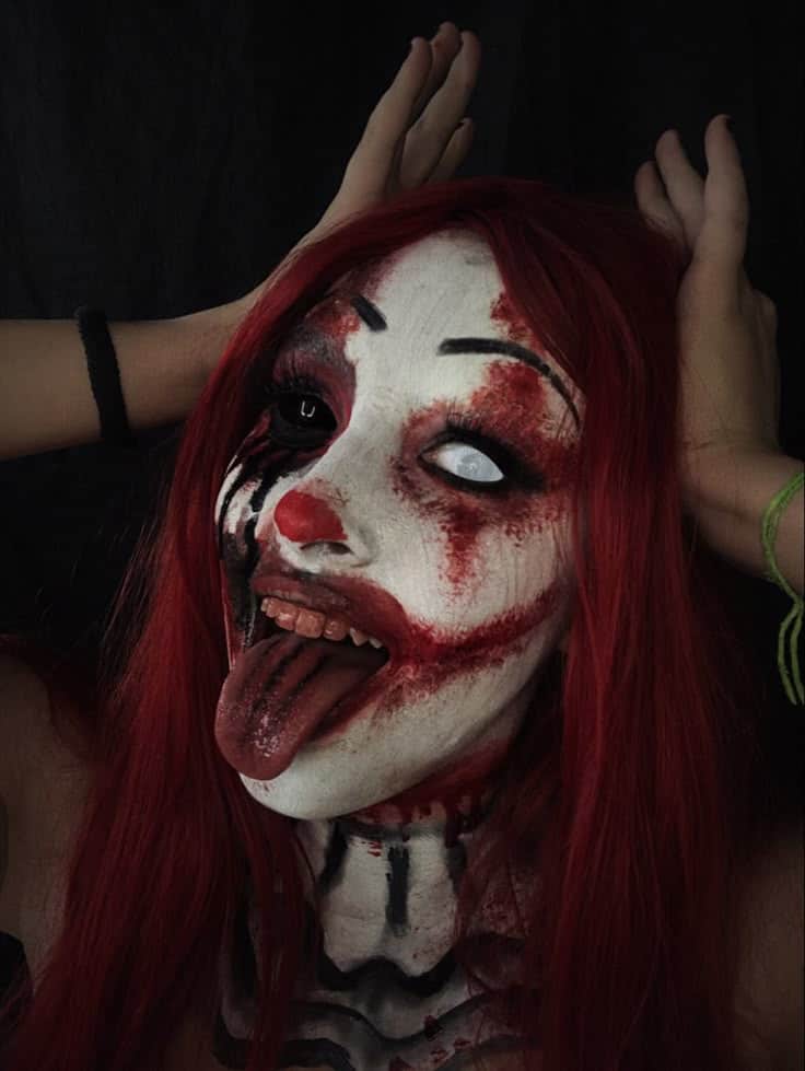 Scary clown with red hair, face paint, and bloody makeup, sticking tongue out against a dark background. Spooky Halloween look.