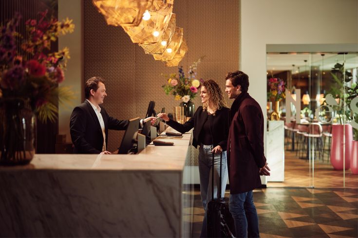 Hotel Hotel guests checking in at reception desk, greeted by a friendly staff member in a modern, warmly lit lobby. | Sky Rye Design Hotel guests checking in at reception desk, greeted by a friendly staff member in a modern, warmly lit lobby.