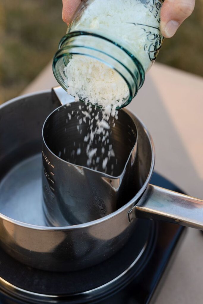 Pouring grated wax from a jar into a metal pitcher for candle making on a stovetop.