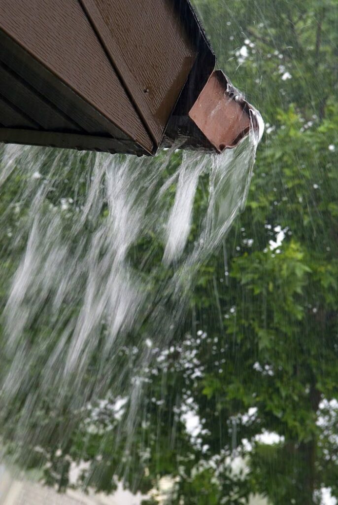 Overflowing gutter during heavy rain, water cascading down with blurred green trees in the background.