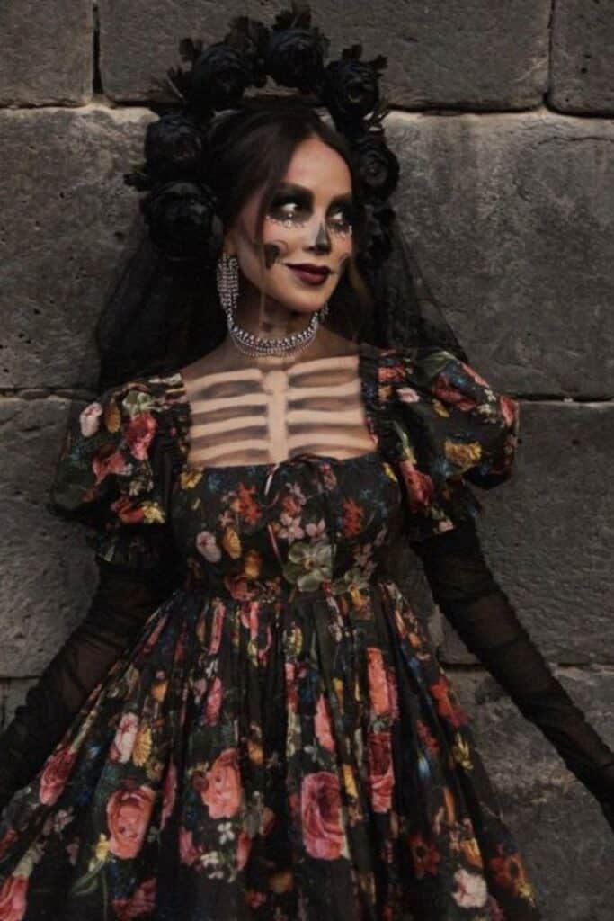Woman in floral dress with skeleton makeup and black floral headpiece during Day of the Dead celebration.