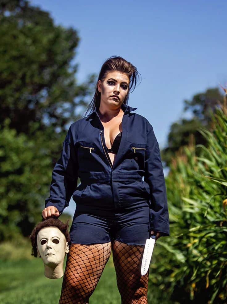 Person in a blue jumpsuit holding a mask and knife, standing in a grassy field under a clear blue sky.