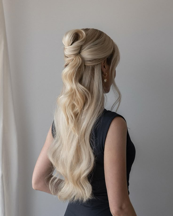 Blonde woman with long, wavy hair in a half-up knot hairstyle, wearing a black dress, facing away.